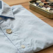 Aiguille et fil traversant un bouton posé sur une chemise en coton bleu clair, avec boîte à boutons ancienne