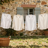 Chemises sur cintres en bois et linge blanc séchant sur un fil dans un jardin ensoleillé, panier en osier posé dans l'herbe